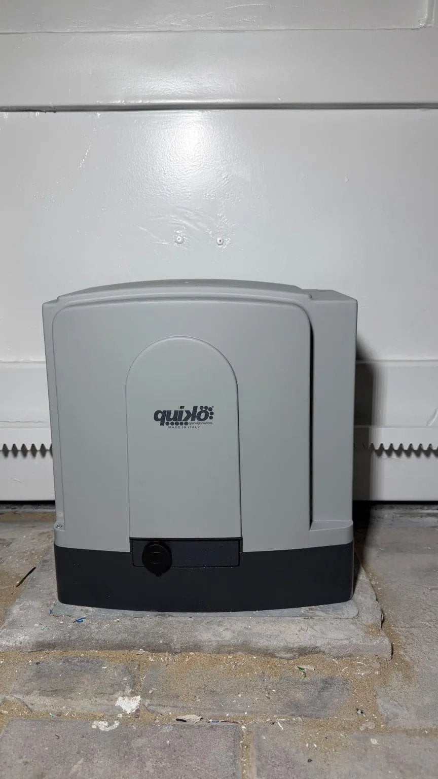 Quiko MOOVY Sliding Gate Operator, gray plastic motor housing with dark base mounted beside a metal gate rack